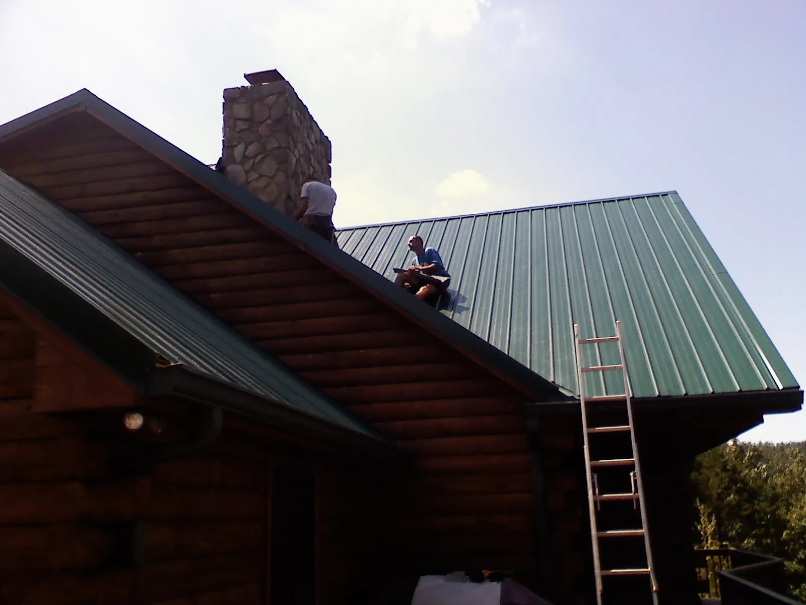 Roofers installing green metal panels beside a stone chimney on a log home in Creston